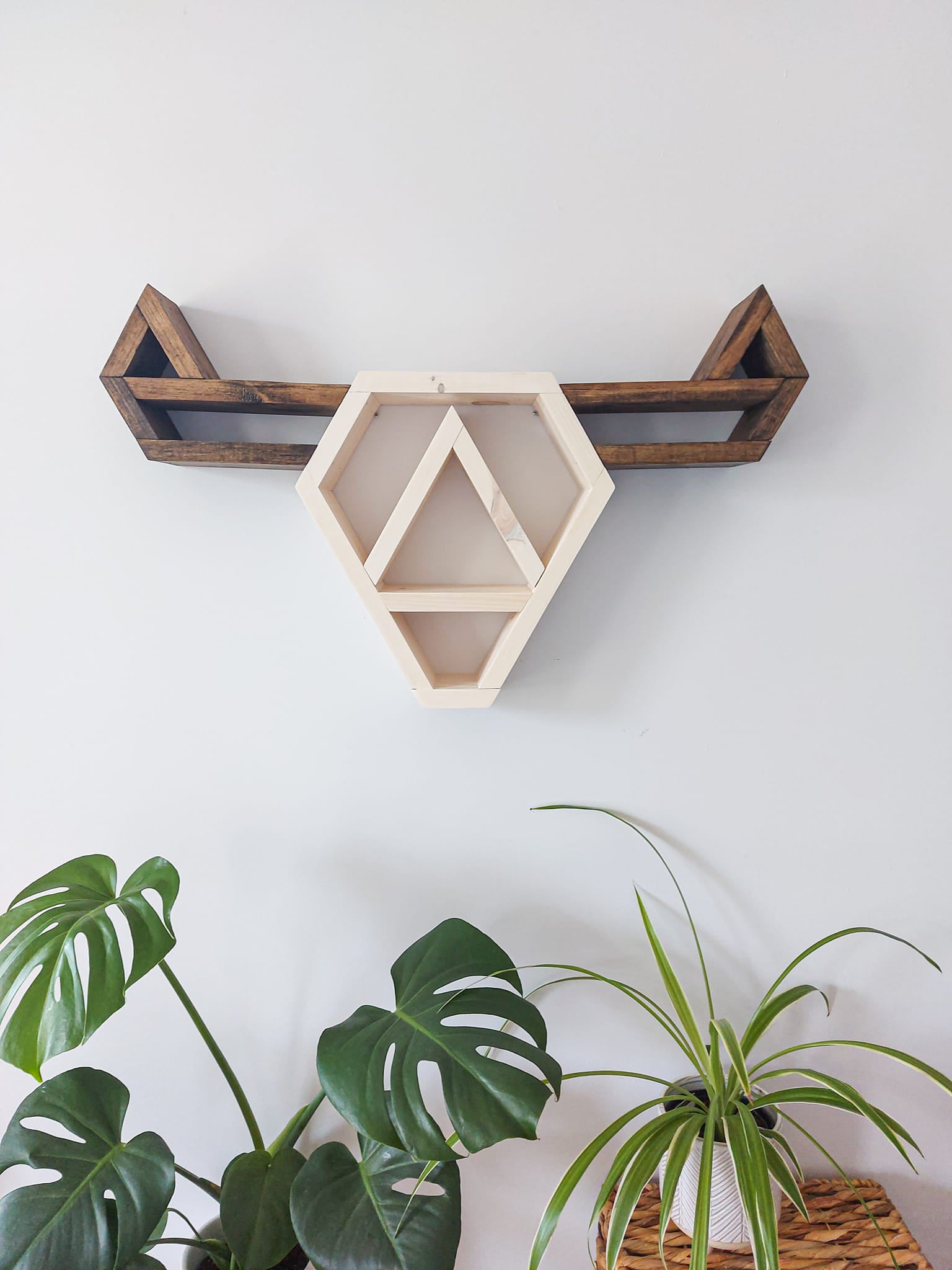 A wooden geometric shelf shaped like a cow head, stained in a light color, mounted on a wall above a wicker basket and green foliage.